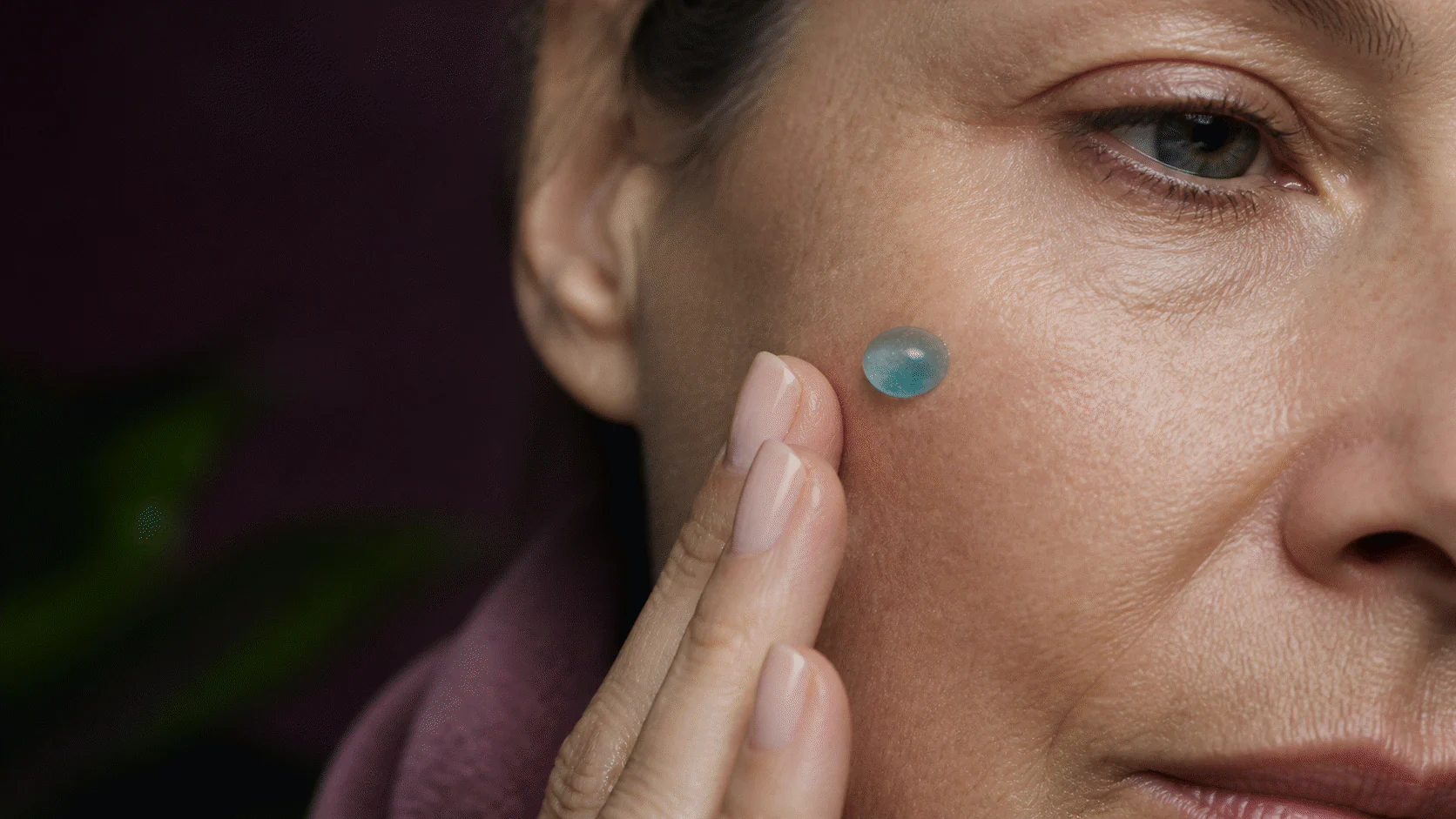 Close view of naturally textured mature cheek skin with a small blue serum droplet and fingertips nearby.