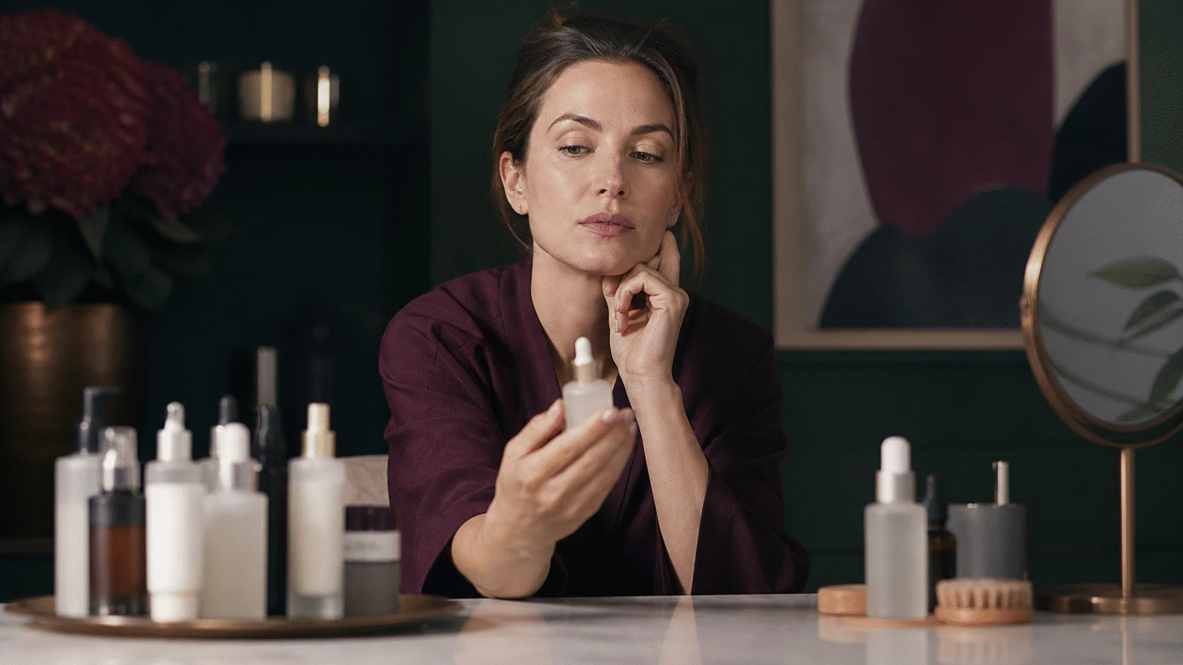 Woman thoughtfully choosing one unlabeled serum among several products at a dark green vanity.