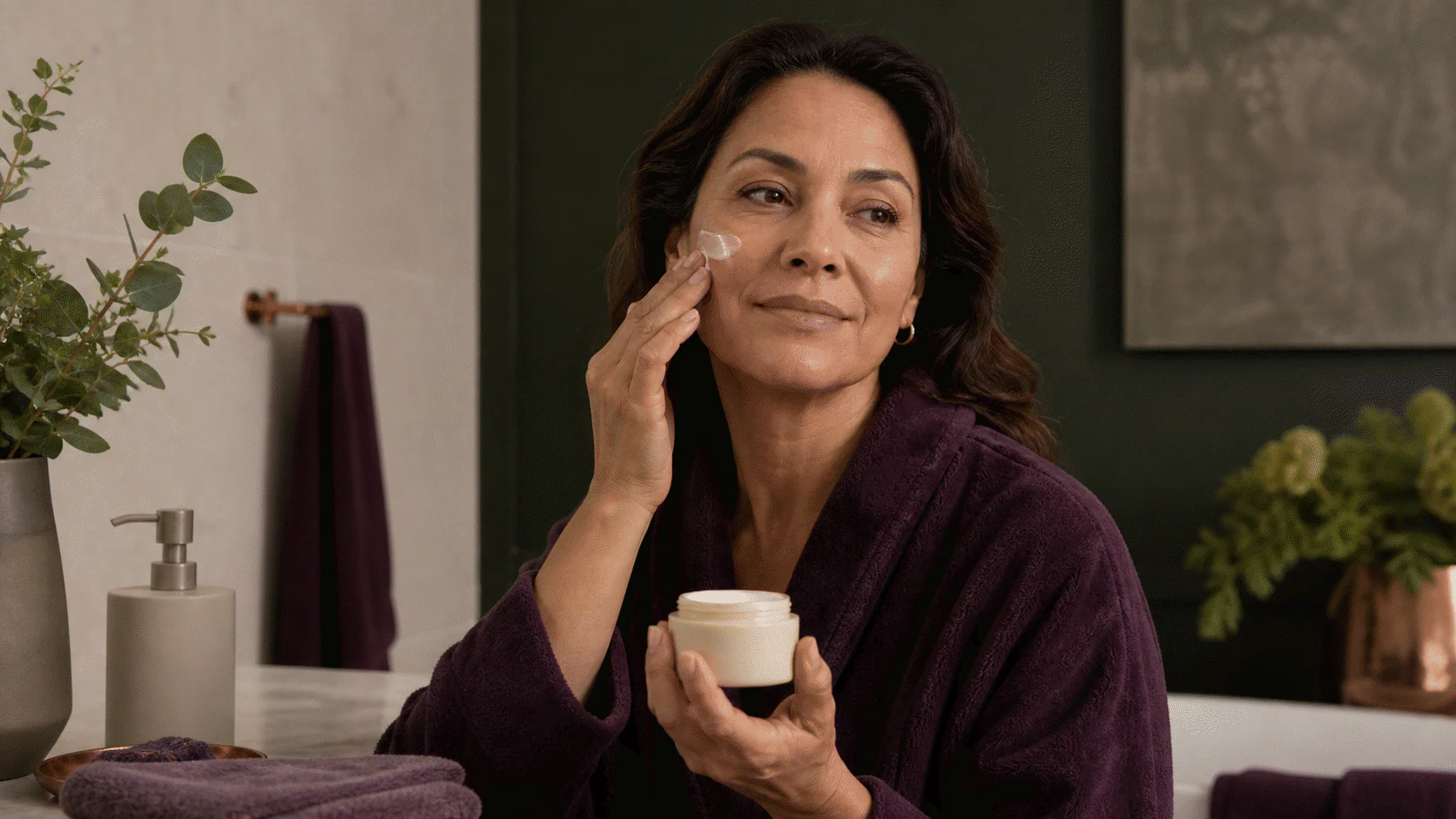 Woman in a plum robe applying a simple cream while holding an unlabeled jar.