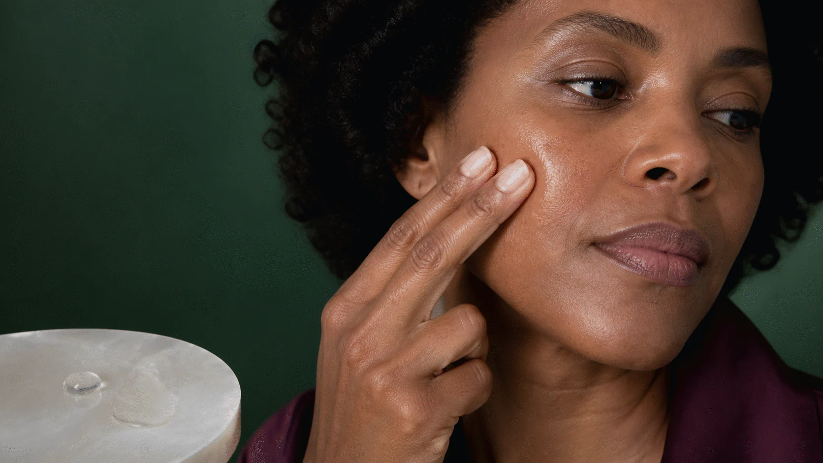 Woman pressing a light facial oil into her cheek beside a clear droplet and thin oil film.