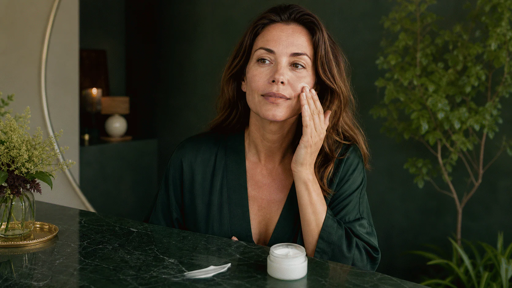 Woman with natural skin texture applying cream to her cheek at a forest-green vanity.