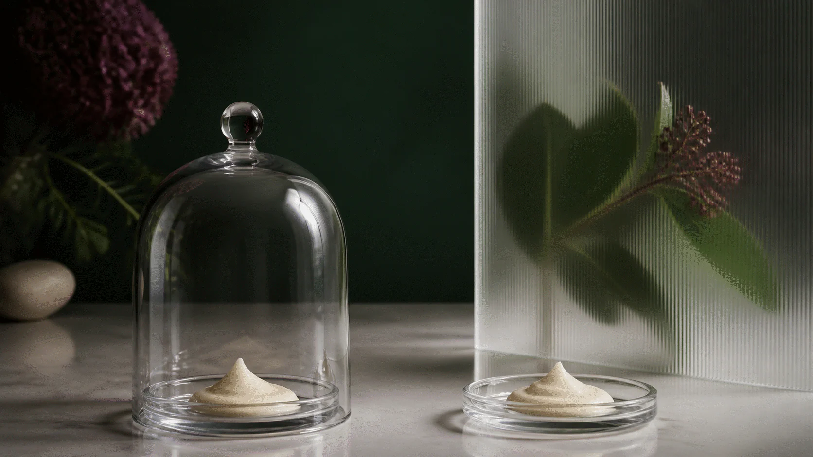 Two matching cream samples, one beneath a clear cloche and one beside a botanical behind frosted glass.
