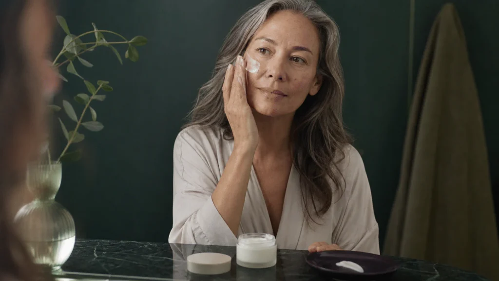 Woman with silver-streaked hair applying a simple cream at a dark green vanity.