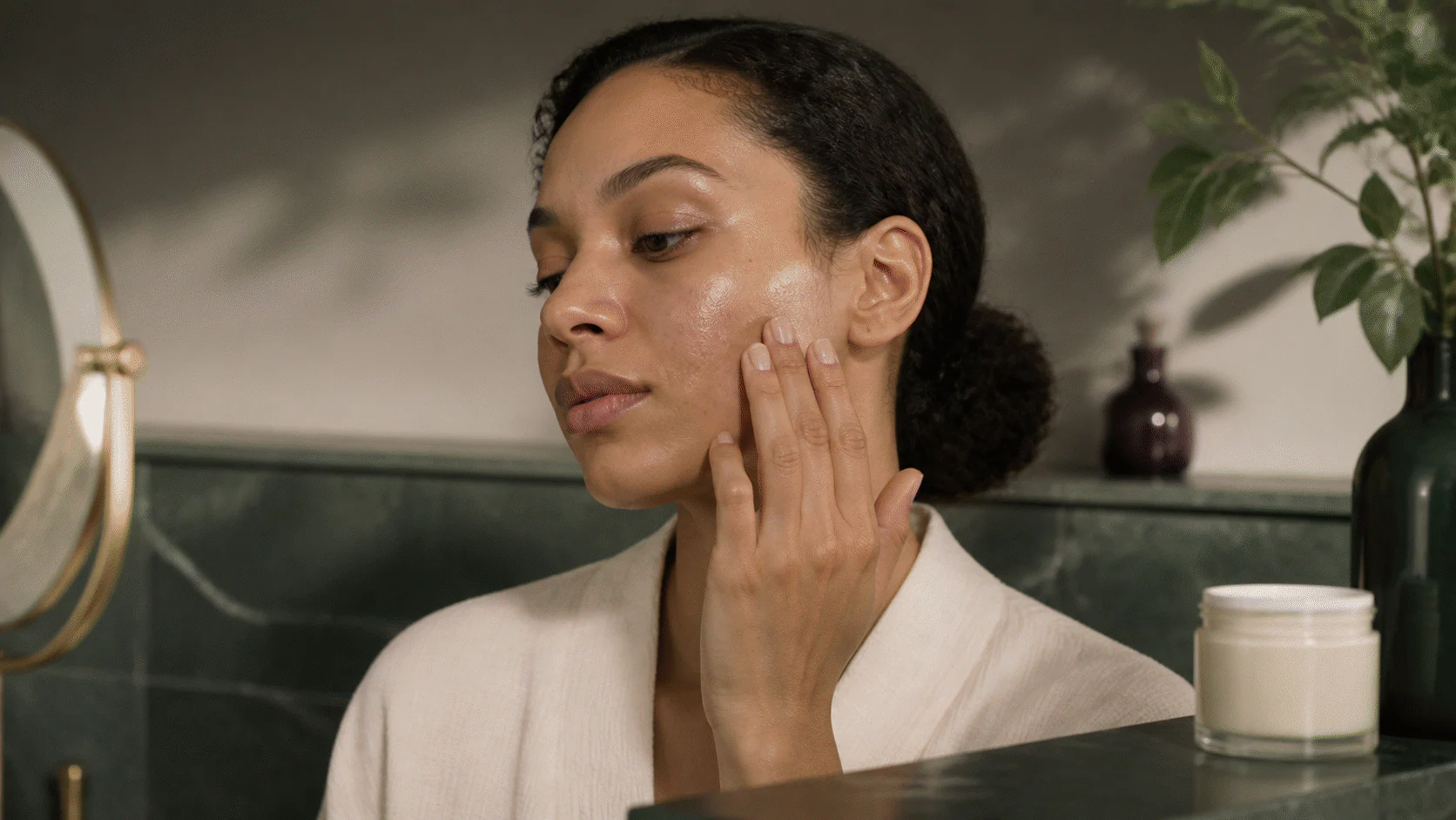 Woman calmly applying skincare to naturally textured cheek skin beside a plain moisturizer jar.