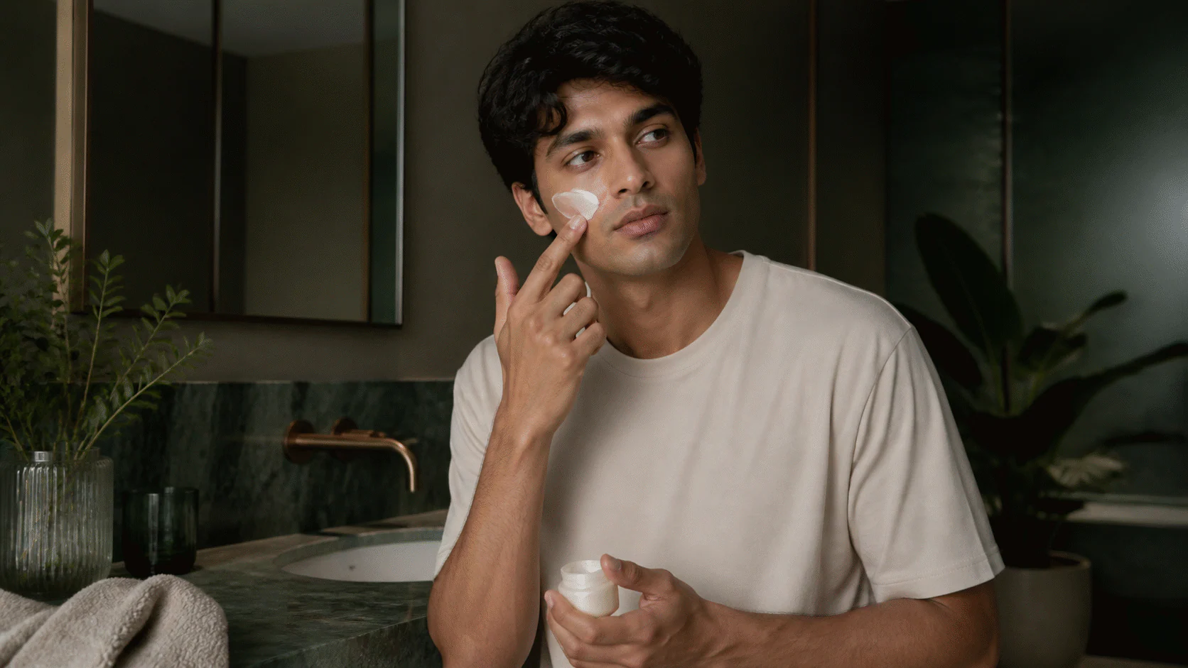 Young man applying pearl cream to his cheek at a bathroom sink with the faucet turned off.