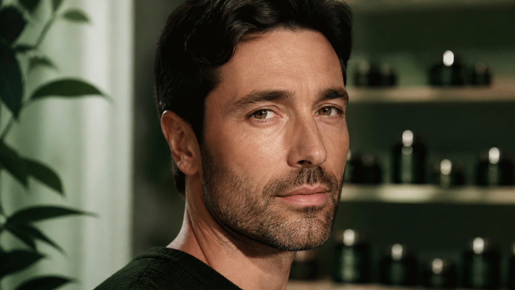 Close portrait of a man with natural cheek texture in front of shelves of blurred unlabelled skincare jars.