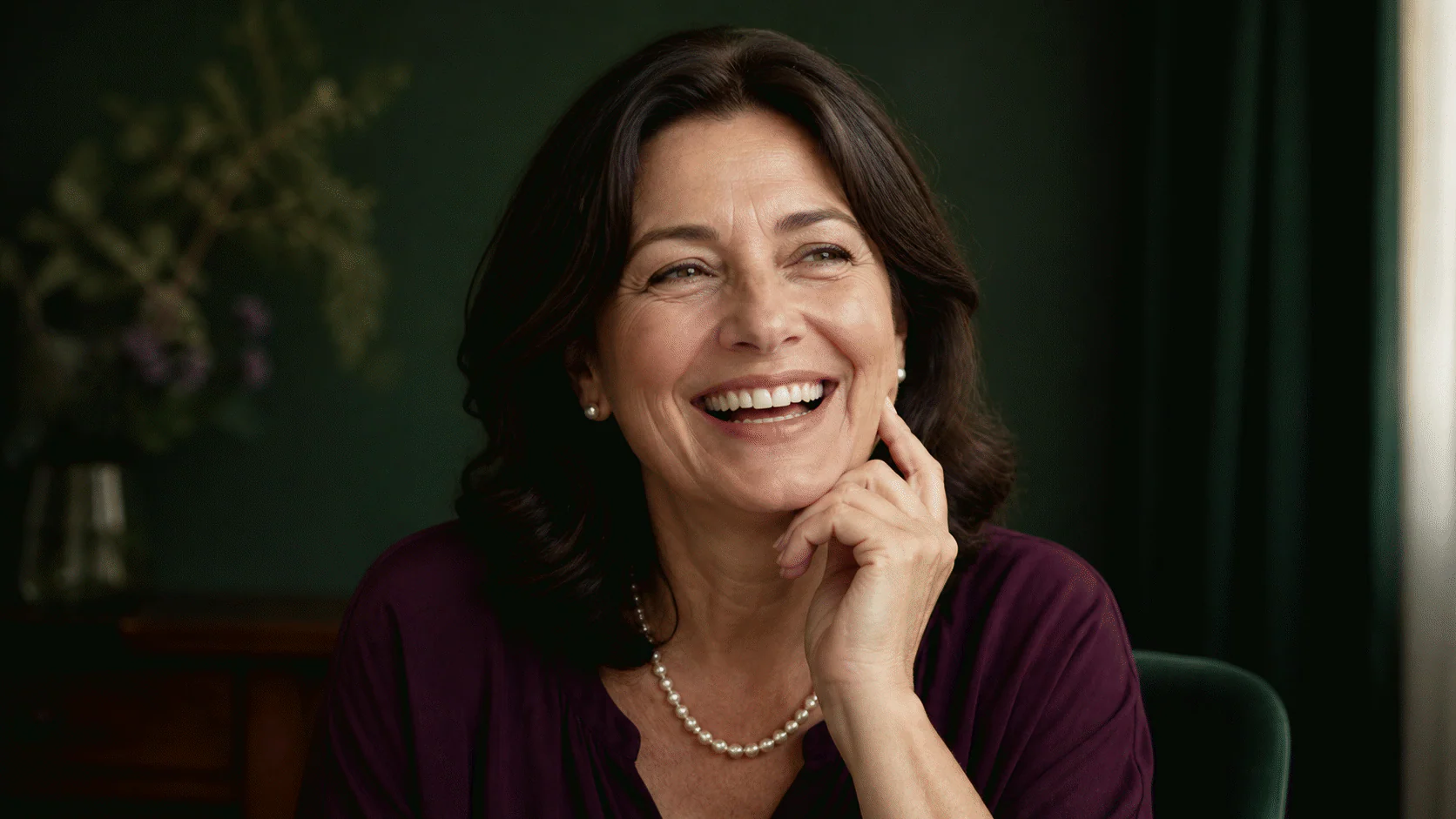 Joyful woman around fifty laughing with natural nasolabial folds visible against a deep green background.